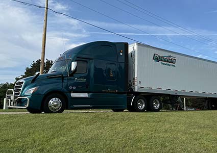 A green Plainfield Trucking semi with a white trailer parked on grass under a blue sky.