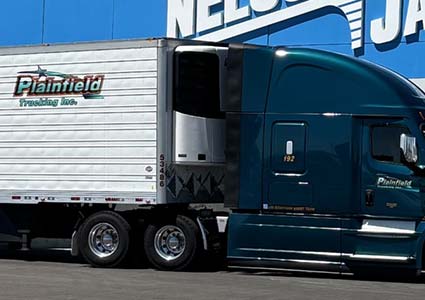 A teal Plainfield Trucking dry van is parked in front of a blue wall with white text.
